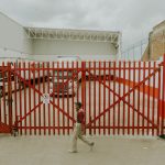 Homem caminha diante de um portão industrial vermelho, com caminhões estacionados dentro de uma instalação logística.