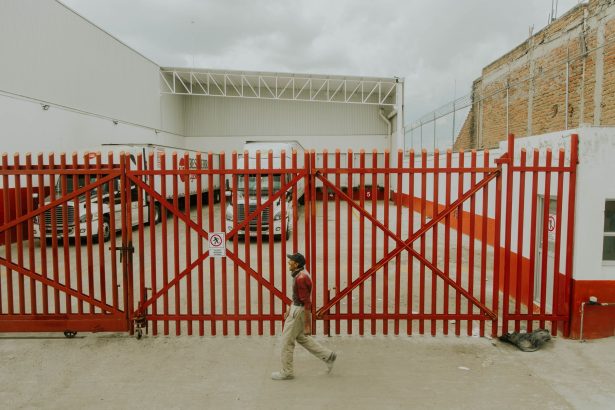 Homem caminha diante de um portão industrial vermelho, com caminhões estacionados dentro de uma instalação logística.