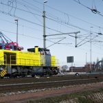 Locomotiva amarela circula entre contêineres e estruturas ferroviárias em uma área portuária.