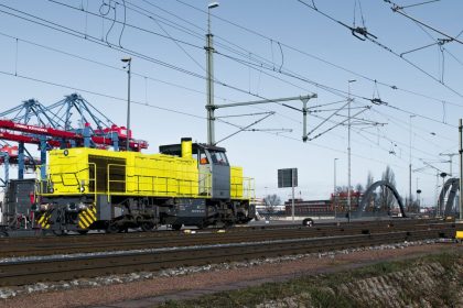 Locomotiva amarela circula entre contêineres e estruturas ferroviárias em uma área portuária.