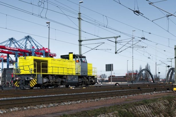 Locomotiva amarela circula entre contêineres e estruturas ferroviárias em uma área portuária.