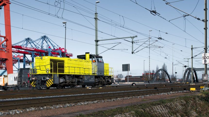 Locomotiva amarela circula entre contêineres e estruturas ferroviárias em uma área portuária.