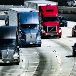 Caminhões e carros trafegam por uma rodovia urbana durante o dia.