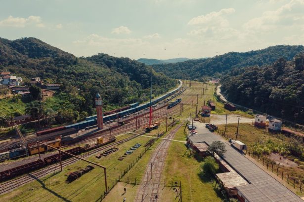 Vista aérea de uma ferrovia atravessando uma paisagem rural cercada por morros verdes.