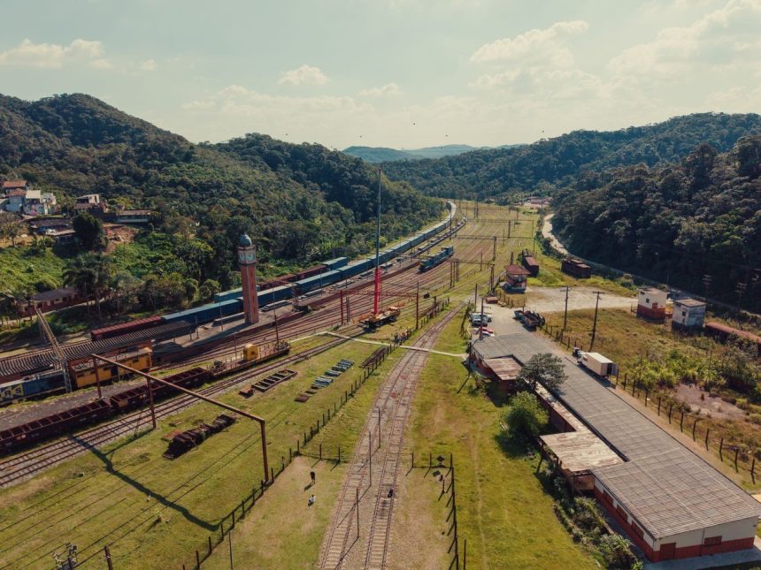 Vista aérea de uma ferrovia atravessando uma paisagem rural cercada por morros verdes.