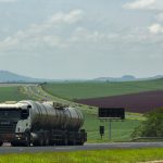 Caminhão de carga trafega por uma rodovia em meio a campos agrícolas.