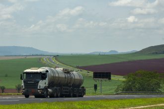 Caminhão de carga trafega por uma rodovia em meio a campos agrícolas.
