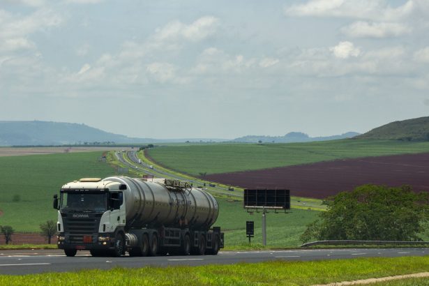 Caminhão de carga trafega por uma rodovia em meio a campos agrícolas.