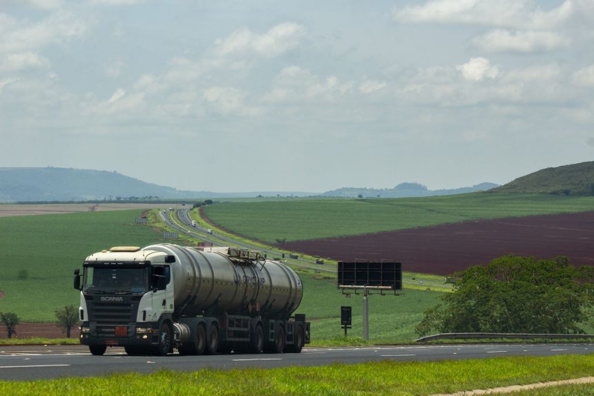 Caminhão de carga trafega por uma rodovia em meio a campos agrícolas.